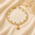 A double-strand gold chain anklet with white faux-pearl beads and a gold seashell charm, displayed on a beige satin fabric background with dried baby's breath flowers.
