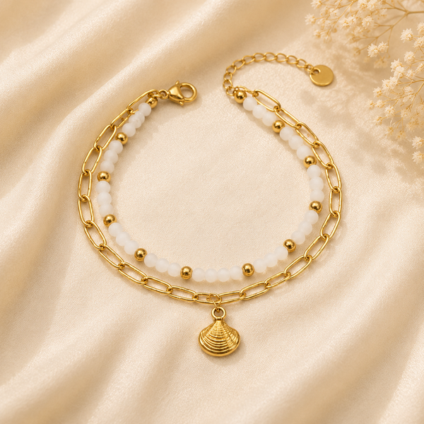 A double-strand gold chain anklet with white faux-pearl beads and a gold seashell charm, displayed on a beige satin fabric background with dried baby's breath flowers.