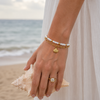 A close-up of a person's hand at the beach wearing layered bracelets and a ring, with a seashell in the foreground and the ocean in the background.