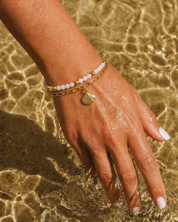 A close-up of a person's hand resting in shallow water, wearing two delicate gold bracelets: one with white beads and one with a small gold seashell charm.